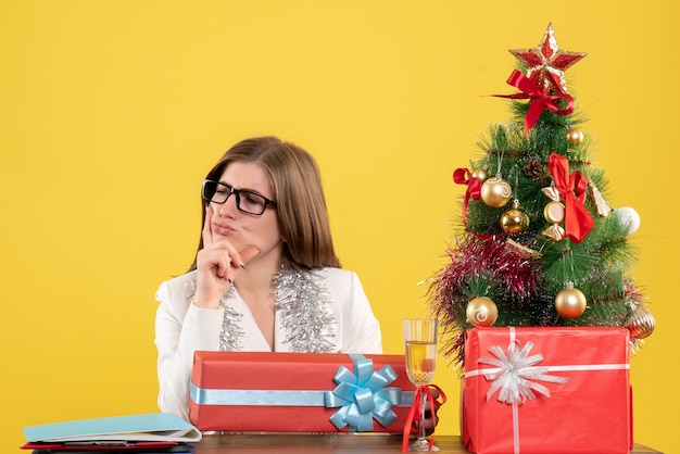 Médica vista frontal sentada em frente à mesa com presentes e árvore em fundo amarelo com árvore de natal e caixas de presente