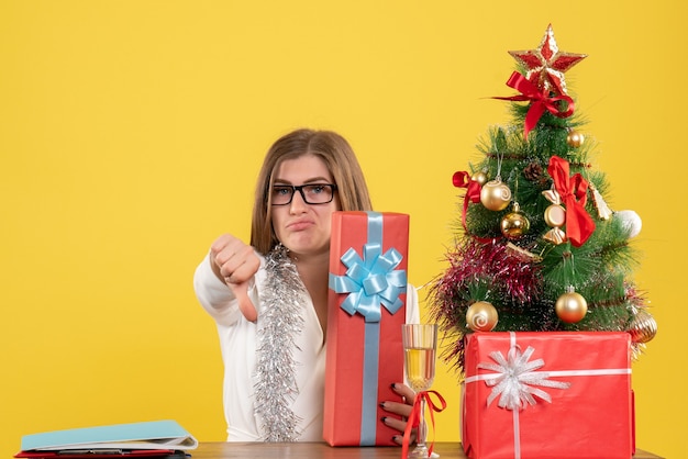 Médica vista frontal sentada em frente à mesa com presentes e árvore em fundo amarelo com árvore de Natal e caixas de presente