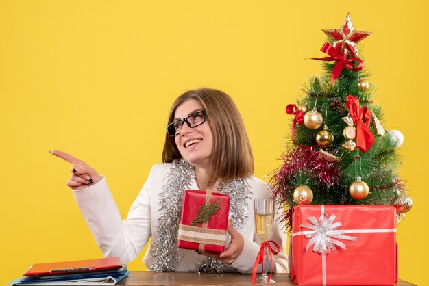 Médica vista frontal sentada em frente à mesa com presentes e árvore em fundo amarelo com árvore de Natal e caixas de presente