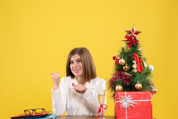 Médica vista frontal sentada em frente à mesa, sorrindo sobre fundo amarelo com árvore de Natal e caixas de presente