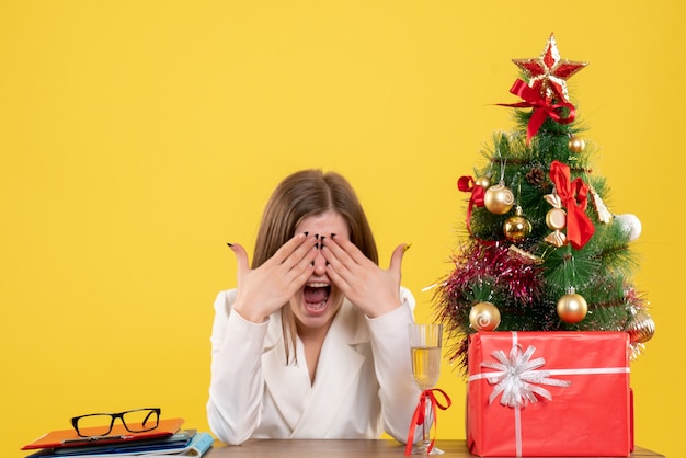 Médica vista frontal sentada em frente à sua mesa em um fundo amarelo com árvore de natal e caixas de presente