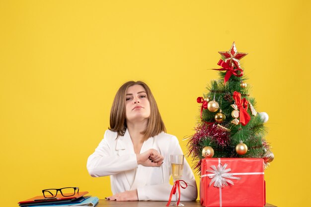 Médica vista frontal sentada em frente à sua mesa em um fundo amarelo com árvore de natal e caixas de presente