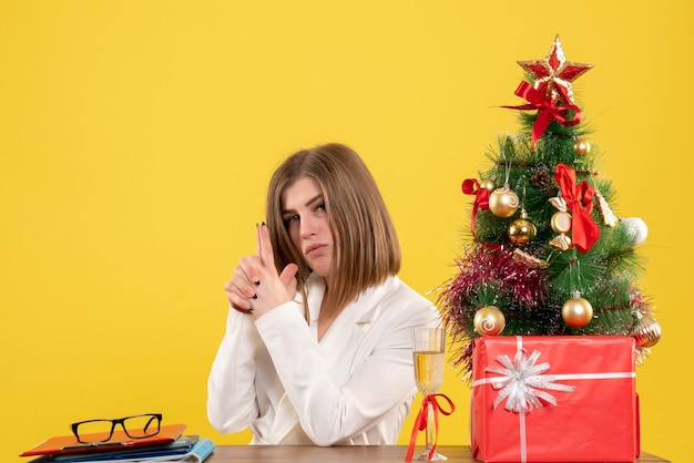Médica vista frontal sentada em frente à sua mesa em um fundo amarelo com árvore de natal e caixas de presente