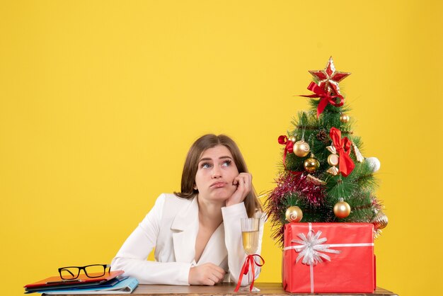 Médica vista frontal sentada em frente à sua mesa em um fundo amarelo com árvore de Natal e caixas de presente