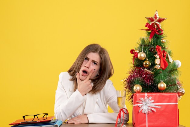 Médica vista frontal sentada em frente à sua mesa em um fundo amarelo com árvore de natal e caixas de presente