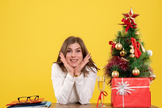 Médica vista frontal sentada em frente à sua mesa em um fundo amarelo com árvore de Natal e caixas de presente