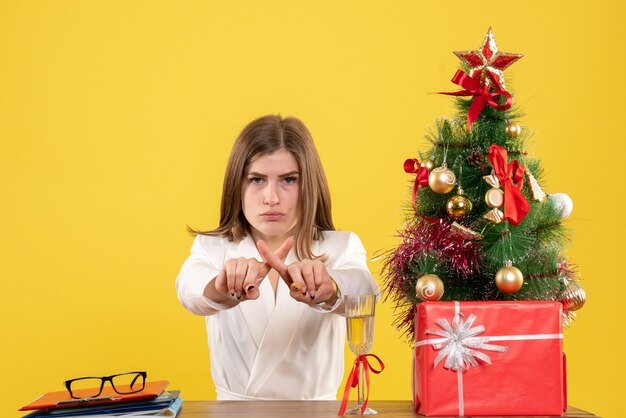 Médica vista frontal sentada em frente à sua mesa em um fundo amarelo com árvore de Natal e caixas de presente