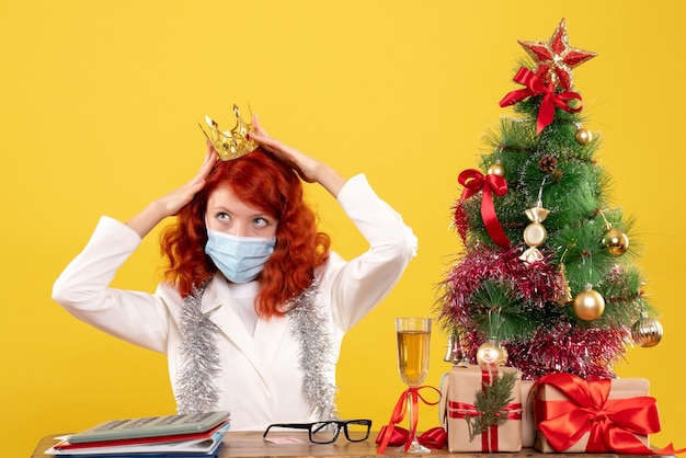 Médica vista frontal sentada na máscara com presentes de natal usando coroa sobre fundo amarelo