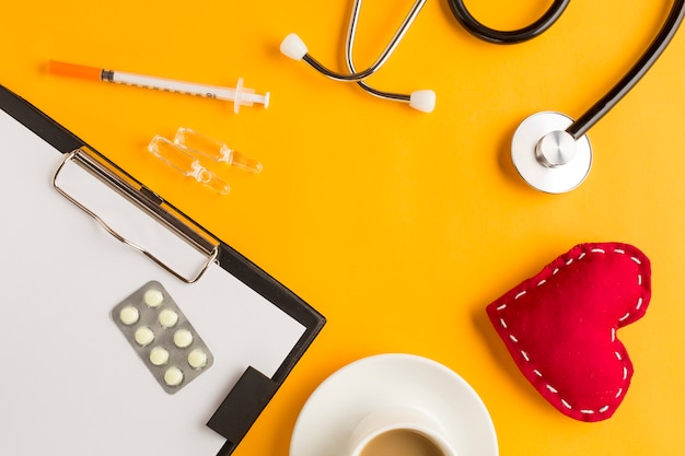 Foto grátis medicamentos sobre a prancheta com coração costurado; ampola; estetoscópio e xícara de café; injeção em fundo amarelo