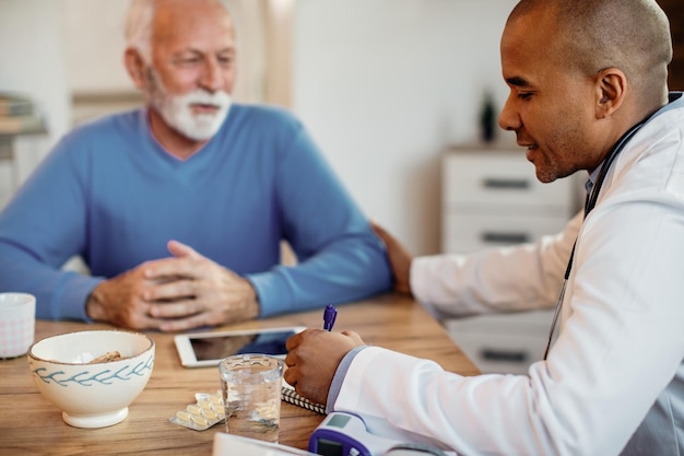 Médico afro-americano tomando notas enquanto conversa com paciente sênior no lar de idosos