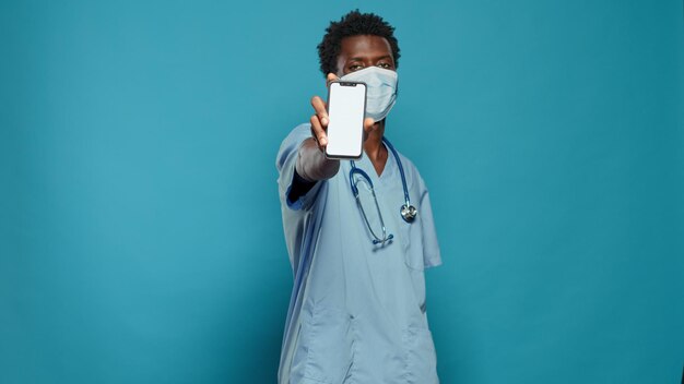 Médico assistente usando máscara facial para proteger contra vírus, mostrando a tela em branco no smartphone. Homem trabalhando como enfermeiro e protegendo contra a pandemia de coronavírus, segurando um visor branco de croma