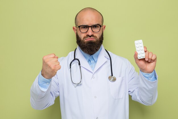 Médico barbudo com jaleco branco com estetoscópio no pescoço e óculos segurando uma bolha com comprimidos, olhando com cara séria mostrando o punho