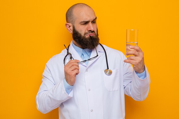 Médico barbudo com jaleco branco com estetoscópio no pescoço segurando um copo d'água olhando para ele com expressão pensativa no rosto pensando em pé sobre um fundo laranja