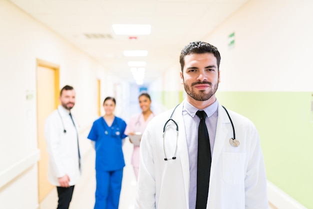 Médico bonito hispânico ao lado da parede com colegas de trabalho em segundo plano no hospital