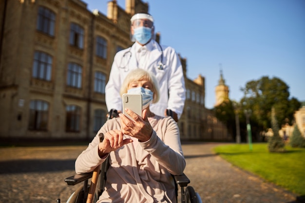 Médico com escudo protetor e jaleco branco empurrando a cadeira de rodas de uma senhora idosa com um smartphone moderno