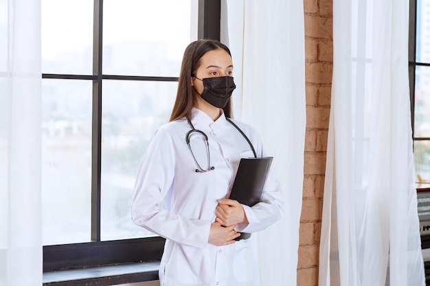 Médico com estetoscópio e máscara preta em pé ao lado da janela e segurando uma pasta preta com o histórico dos pacientes.