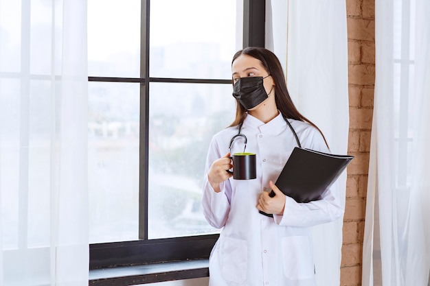 Médico com estetoscópio e máscara preta segurando um copo preto de bebida e uma pasta preta e olhando pela janela.