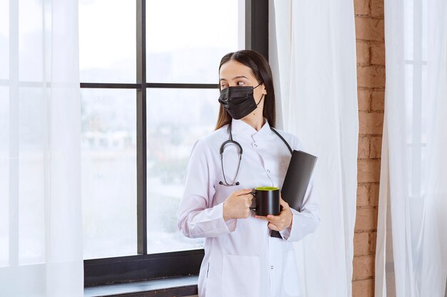 Médico com estetoscópio e máscara preta segurando um copo preto de bebida e uma pasta preta e olhando pela janela.