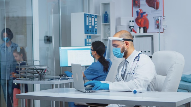 Médico com máscara de proteção e viseira digitando o tratamento no laptop enquanto a mãe acompanha a filha à consulta no hospital durante a pandemia de coronavírus. Enfermeira equipada falando com os pacientes.