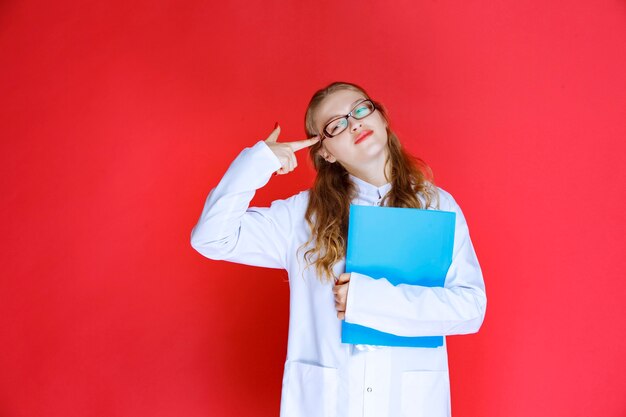 Médico com óculos, segurando uma pasta azul e parece cansado.