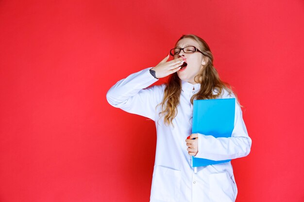 Médico com óculos, segurando uma pasta azul e parece cansado.