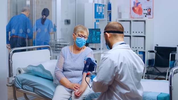 Médico com roupas de proteção, verificando paciente com hipertensão em um hospital ou clínica privada moderna durante a pandemia de COVID-19. Verificação de saúde, diagnóstico de exame médico de doença