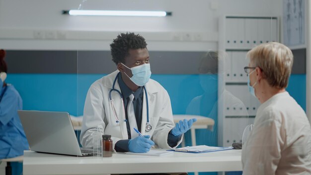 Médico consultando paciente idoso com máscara facial e luvas para proteção contra coronavírus em estabelecimento de saúde. Jovem médico ajudando idosa durante a pandemia na consulta de check-up