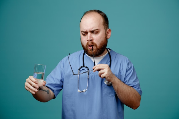 Médico de homem barbudo de uniforme com estetoscópio no pescoço usando óculos segurando um copo de água olhando de perto com cara séria em pé sobre fundo azul