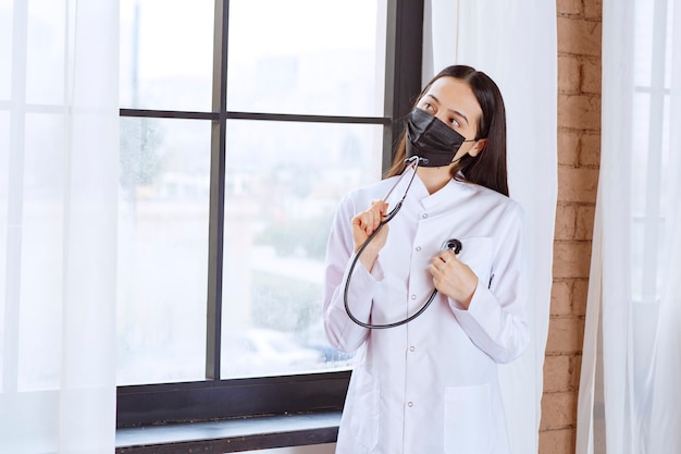 Médico de máscara preta com um estetoscópio em pé perto da janela e verificando os batimentos cardíacos.