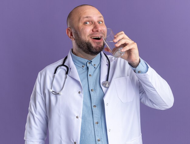 Médico de meia-idade alegre, vestindo túnica médica e estetoscópio segurando um copo de água perto da boca, preparando-se para beber água