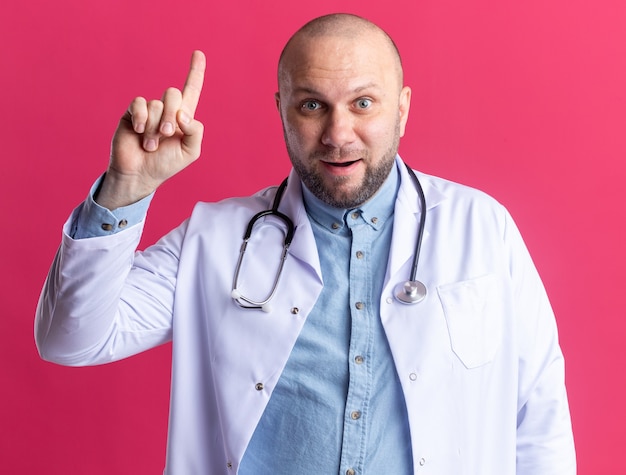 Médico de meia-idade impressionado usando túnica médica e estetoscópio apontando para cima, isolado na parede rosa