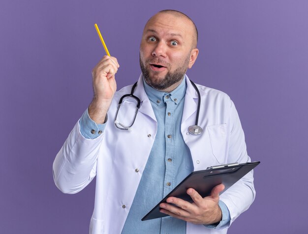 Médico de meia-idade impressionado, vestindo túnica médica e estetoscópio segurando uma prancheta, olhando para a frente, apontando para cima com um lápis isolado na parede roxa