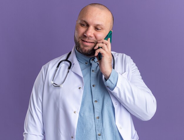 Médico de meia-idade satisfeito, vestindo túnica médica e estetoscópio falando ao telefone, olhando para baixo, isolado na parede roxa