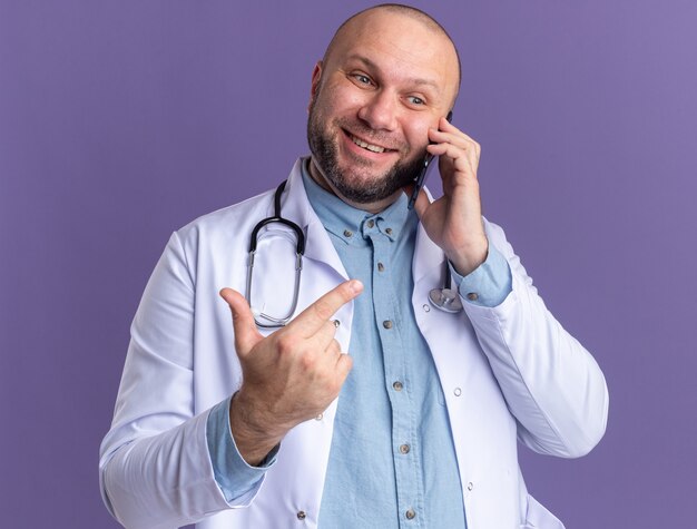 Médico de meia-idade sorridente, vestindo bata médica e estetoscópio falando no telefone, olhando para o lado apontando para o lado isolado na parede roxa