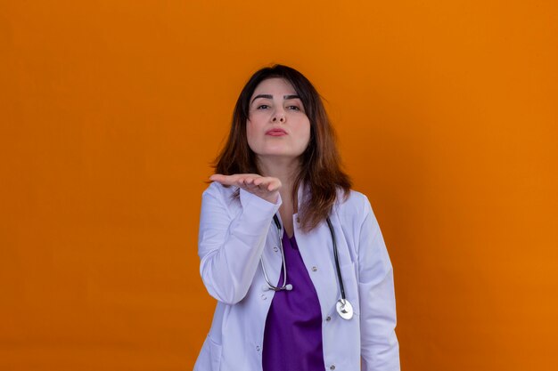 Médico de meia idade vestindo jaleco branco e com estetoscópio, olhando para a câmera mandando um beijo com a mão no ar, sendo adorável sobre parede laranja isolada