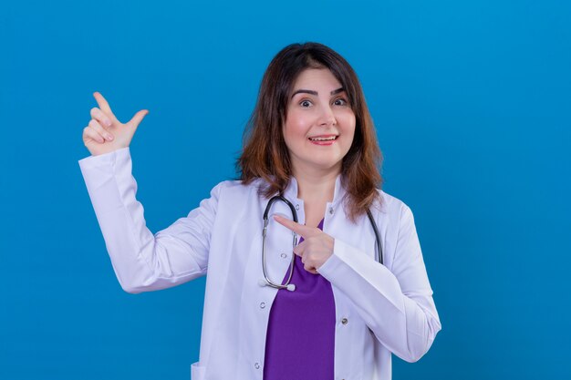 Médico de meia idade vestindo jaleco branco e com estetoscópio sorrindo alegremente apontando com as mãos e os dedos para o lado ao longo da parede azul