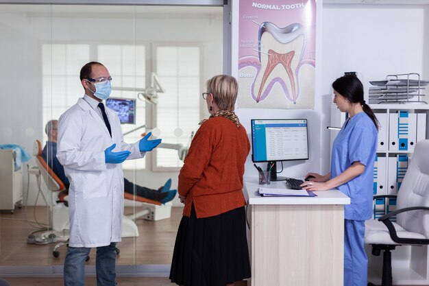 Médico dentista com máscara explicando o diagnóstico para paciente sênior no corredor da área de espera de Estomatologia. Homem idoso sentado na cadeira para tratamento de dentes.