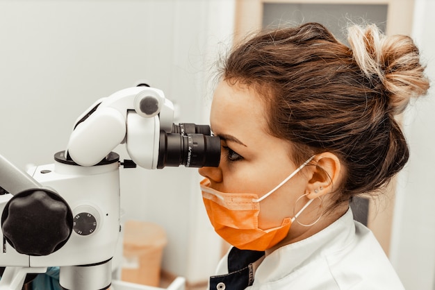 Médico dentista jovem olha através de um microscópio profissional em uma clínica odontológica. um médico em uma máscara médica descartável e boné. equipamento avançado em odontologia