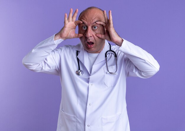 Médico do sexo masculino de meia-idade surpreso, vestindo bata médica e estetoscópio, olhando para a câmera, fazendo grandes olhos isolados no fundo roxo