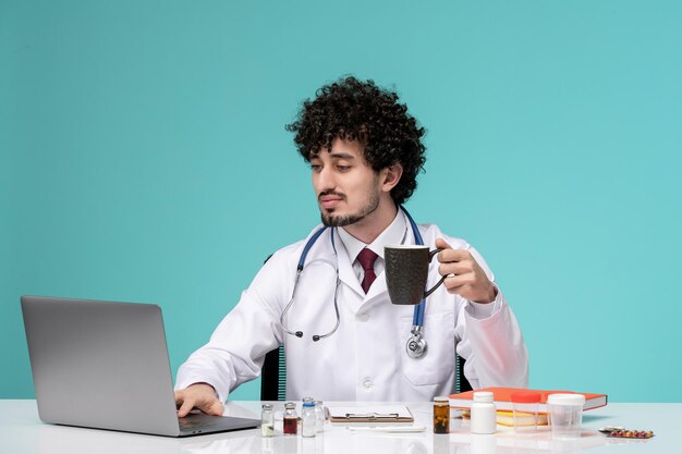 Médico em jaleco jovem médico bonito sério trabalhando no computador segurando uma xícara de café preto