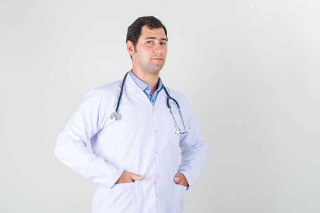 Médico em pé com as mãos nos bolsos, com jaleco branco e parecendo confiante. vista frontal.