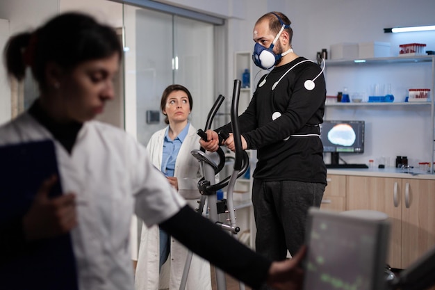 Foto grátis médico especialista olhando para monitor que analisa a pulsação cardíaca enquanto homem atleta correndo no ginásio bycle trabalhando na resistência do corpo durante o treino médico em laboratório moderno. médico monitorando dados de ekg