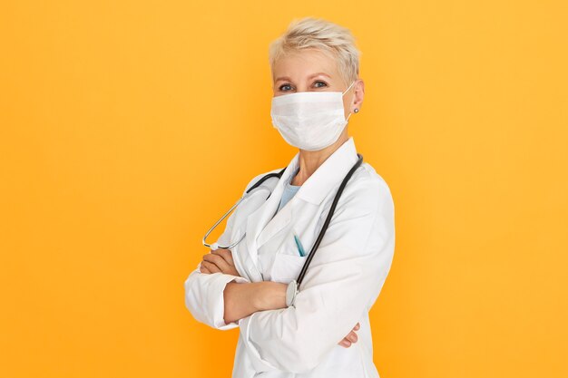 Médico feminino maduro confiante posando contra o fundo da parede amarela em branco, vestindo bata branca médica e máscara cirúrgica protetora, cruzando os braços sobre o peito. Vírus, infecções e bactérias