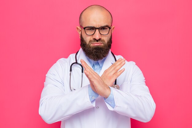 Médico homem barbudo com jaleco branco com estetoscópio no pescoço usando óculos olhando para a câmera com cara séria fazendo gesto de parada cruzando as mãos em pé sobre fundo rosa