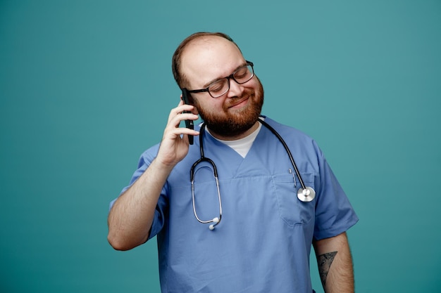 Médico homem barbudo de uniforme com estetoscópio no pescoço usando óculos feliz e satisfeito enquanto fala no celular sorrindo em pé sobre fundo azul