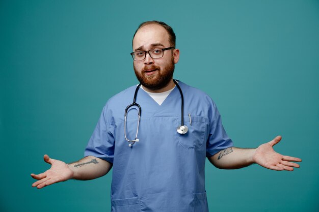 Médico homem barbudo de uniforme com estetoscópio no pescoço usando óculos olhando para a câmera confuso espalhando os braços para os lados sem resposta em pé sobre fundo azul