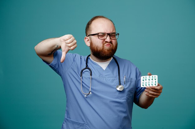 Médico homem barbudo de uniforme com estetoscópio no pescoço usando óculos segurando pílulas olhando para câmera fazendo boca torta descontente mostrando o polegar para baixo em pé sobre fundo azul