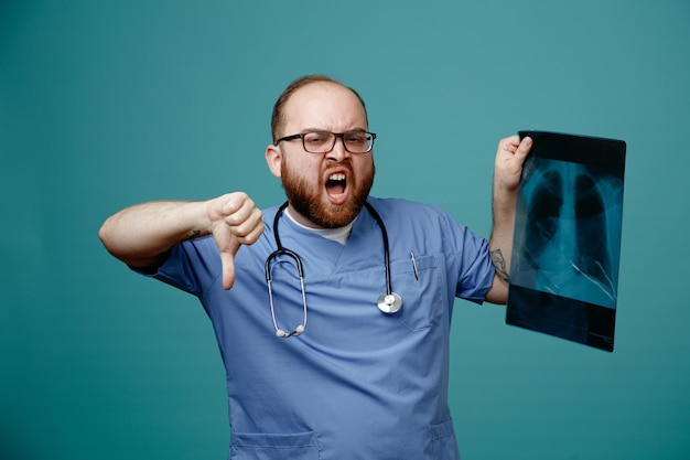 Foto grátis médico homem barbudo de uniforme com estetoscópio no pescoço usando óculos segurando raio-x do pulmão gritando com expressão agressiva, mostrando o polegar para baixo em pé sobre fundo azul