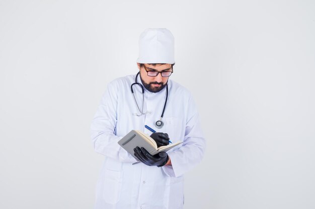 Médico homem de uniforme branco, óculos escrevendo e olhando cuidadoso, vista frontal.