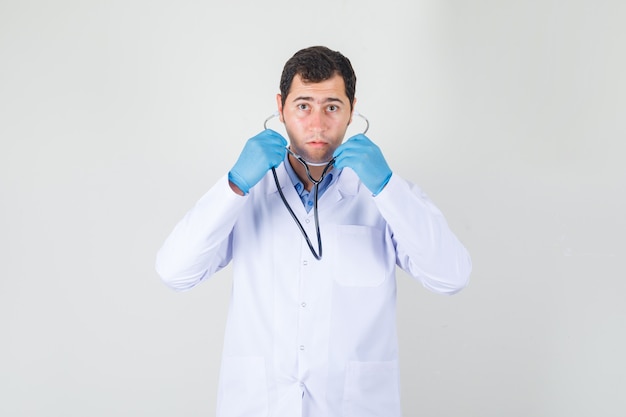 Médico homem usando estetoscópio no jaleco branco, luvas e olhando o foco. vista frontal.